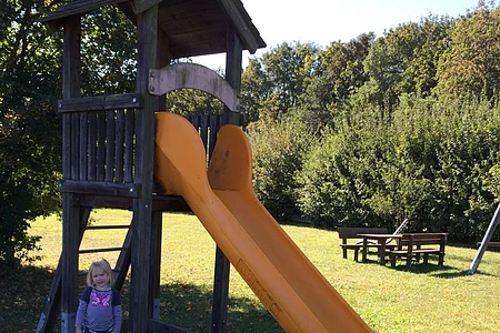Neben der gelben Rutsche lässt sich das Mädchen fotografieren sowie im Hintergrund 2 Holzbänke stehen.