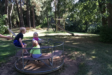 Geschwister sitzen im Rondell, der Spielplatz ist im Wald und im Hintergrund steht eine Rutsche.