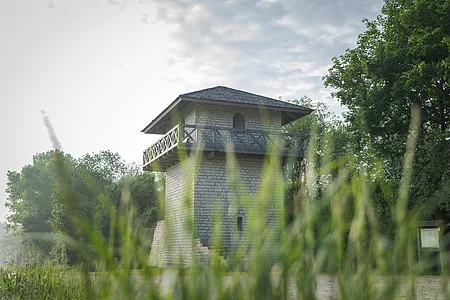 Der Limesturm ist aus weißem Stein rekonstruiert. Die Balustrade ist aus Holz und führt einmal um den ganzen Turm. Es ist unten ein kleines Fenster und oben eine Tür, die auf die Balustrade führt zu sehen. Umrahmt wird der Turm vom grünen Gras und den Bäumen im Hintergrund. Der blaue Himmel ist bewölkt.