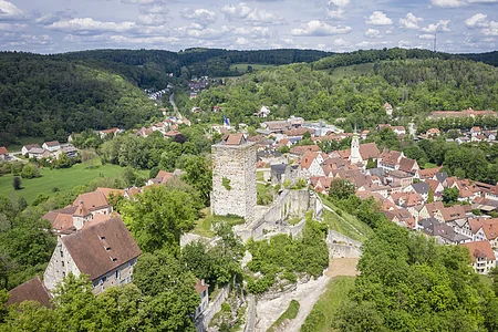 Luftaufnahme einer Burg mit Turm und umliegendem Dorf in einer grünen Hügellandschaft unter bewölktem Himmel.