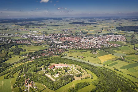 Die Wülzburg mit der gesamten Burganlage von oben. Sie ist komplett umgeben von grünen Gras und Bäumen. Dahinter kann man die Stadt Weißenburg erkennen und den himmelblauen Himmel.