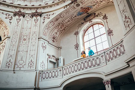Zwei Personen stehen auf einem verzierten Balkon in einem historischen, mit Fresken geschmückten Innenraum.