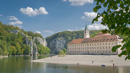 Kloster Weltenburg am Donaudurchbruch Flussufer mit Kiesstrand, altes Gebäude mit Turm und bewaldete Felsen unter blauem Himmel mit Wolken